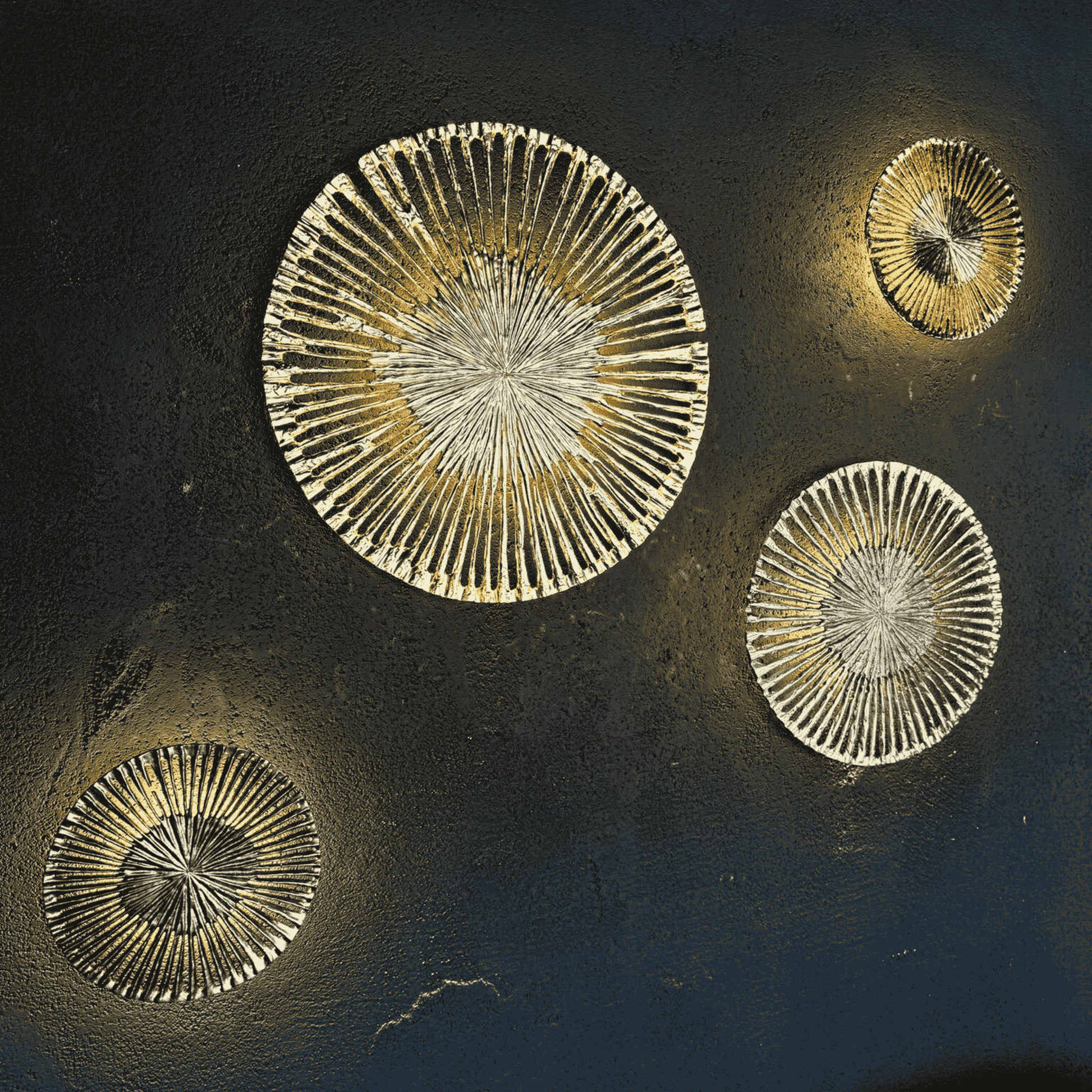 Set of four round cast aluminum wall sconces in sunburst design with gold, nickel, aged brass and black finishes on a dark textured wall