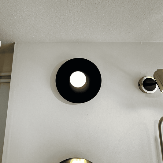 Angled view of black disc wall sconce casting ambient light against a clean white wall.