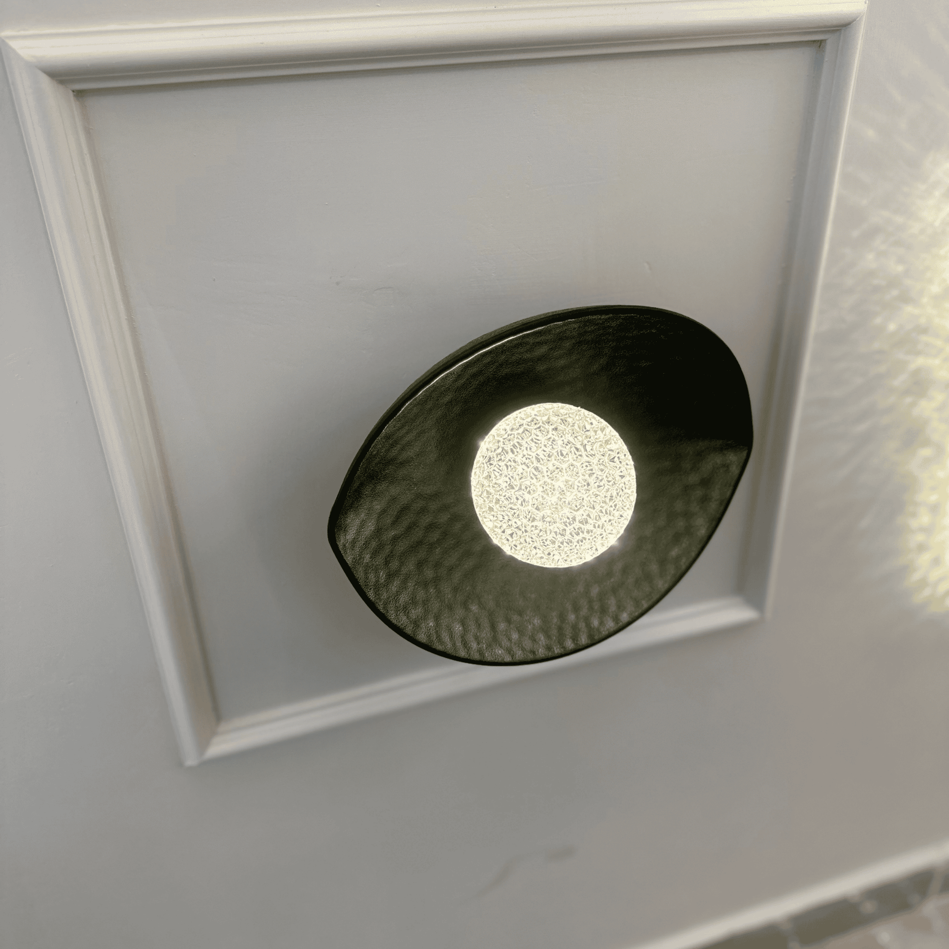 A black plexiglass LED light sconce with a central textured diffuser softly illuminates a neutral-toned wall.