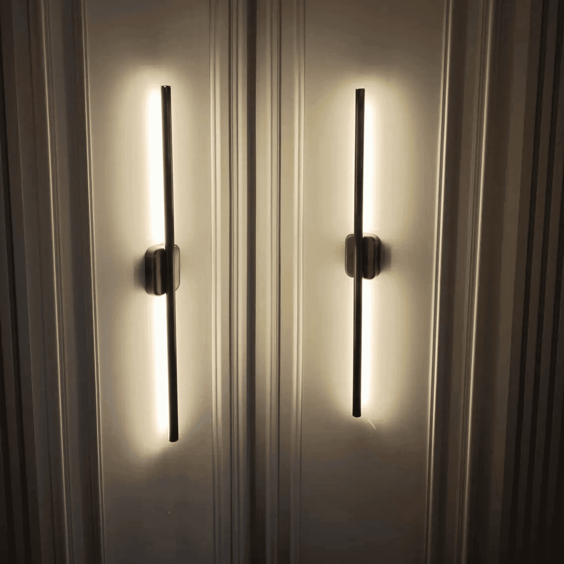 Close-up of two black vertical wall sconces with warm up-and-down light on a decorative paneled wall.