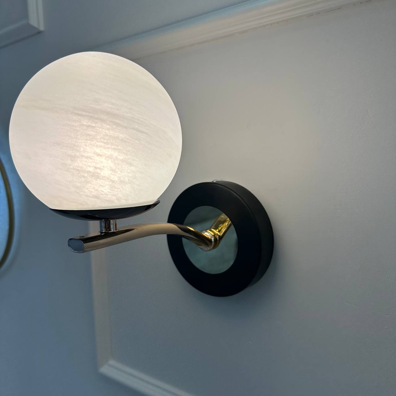 Side angle of a modern alabaster sconce with curved brass arm and black base on a white paneled wall.