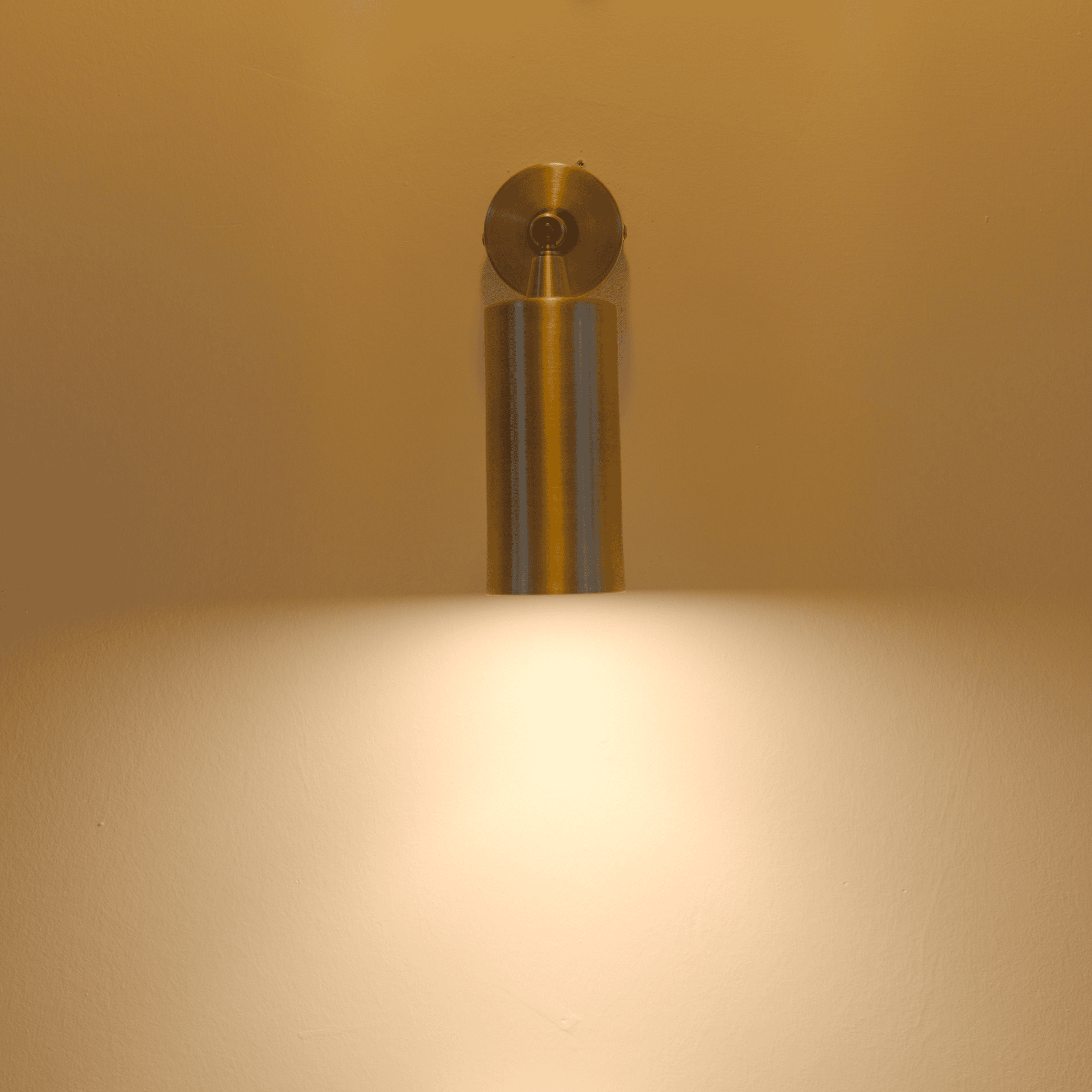 Brass cylindrical wall sconce emitting warm downward light on a neutral wall for modern ambient lighting.