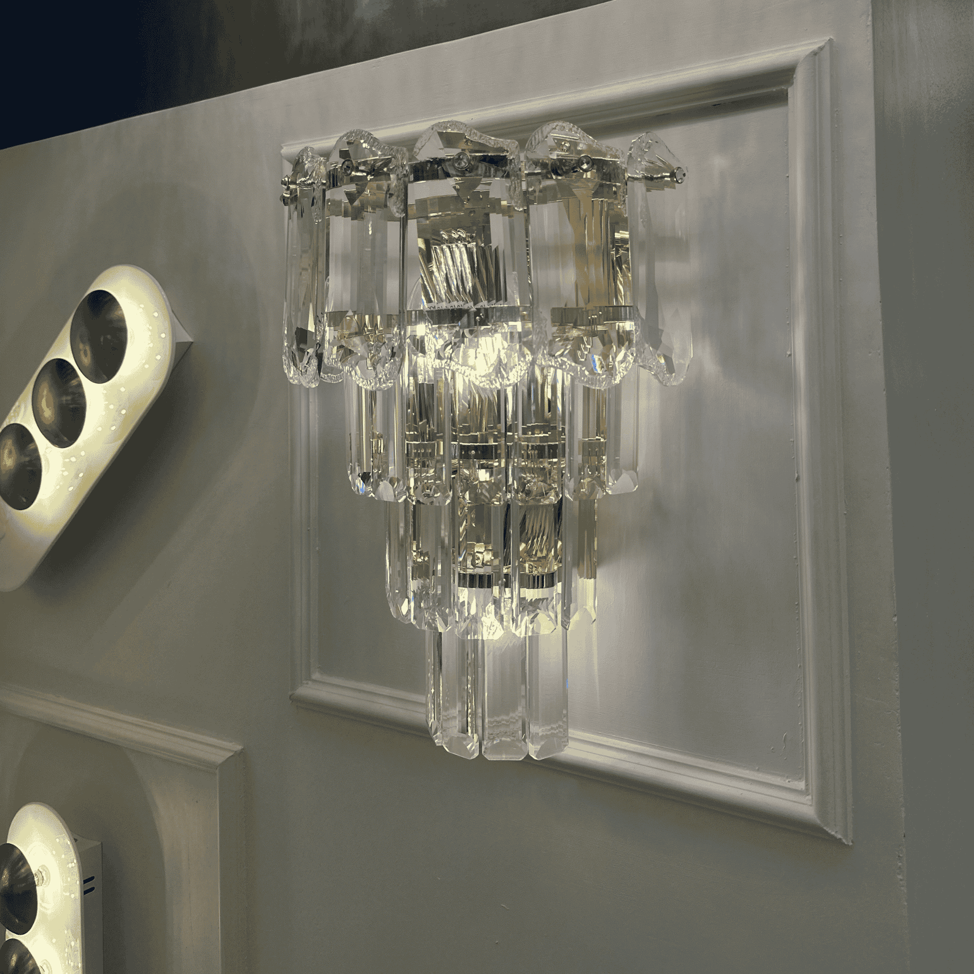 Crystal wall sconce light with layered clear prisms and chrome accents mounted on a wall panel.