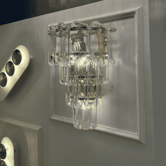 Crystal wall sconce light with layered clear prisms and chrome accents mounted on a wall panel.