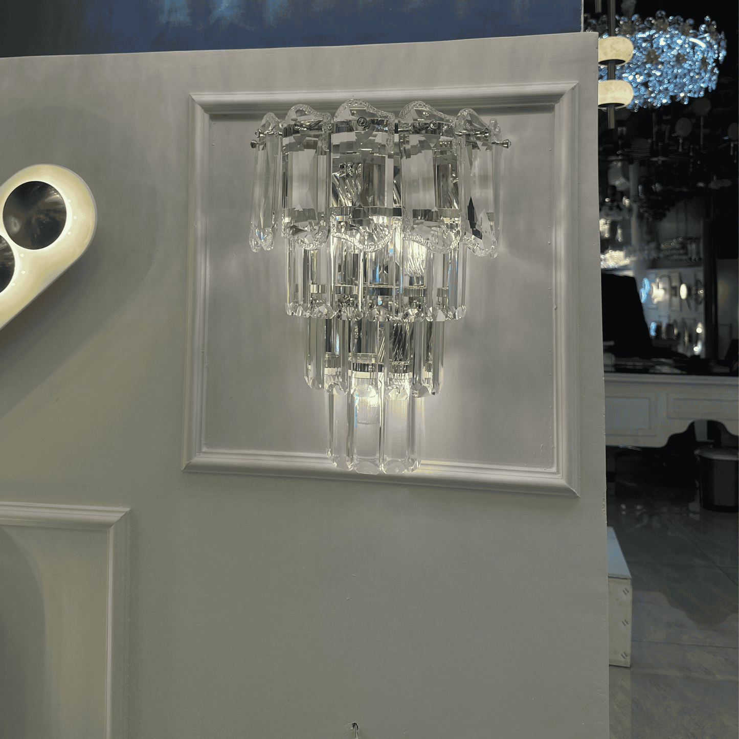 Tiered crystal wall sconce light with elongated clear glass prisms and a chrome frame, mounted on a decorative panel.