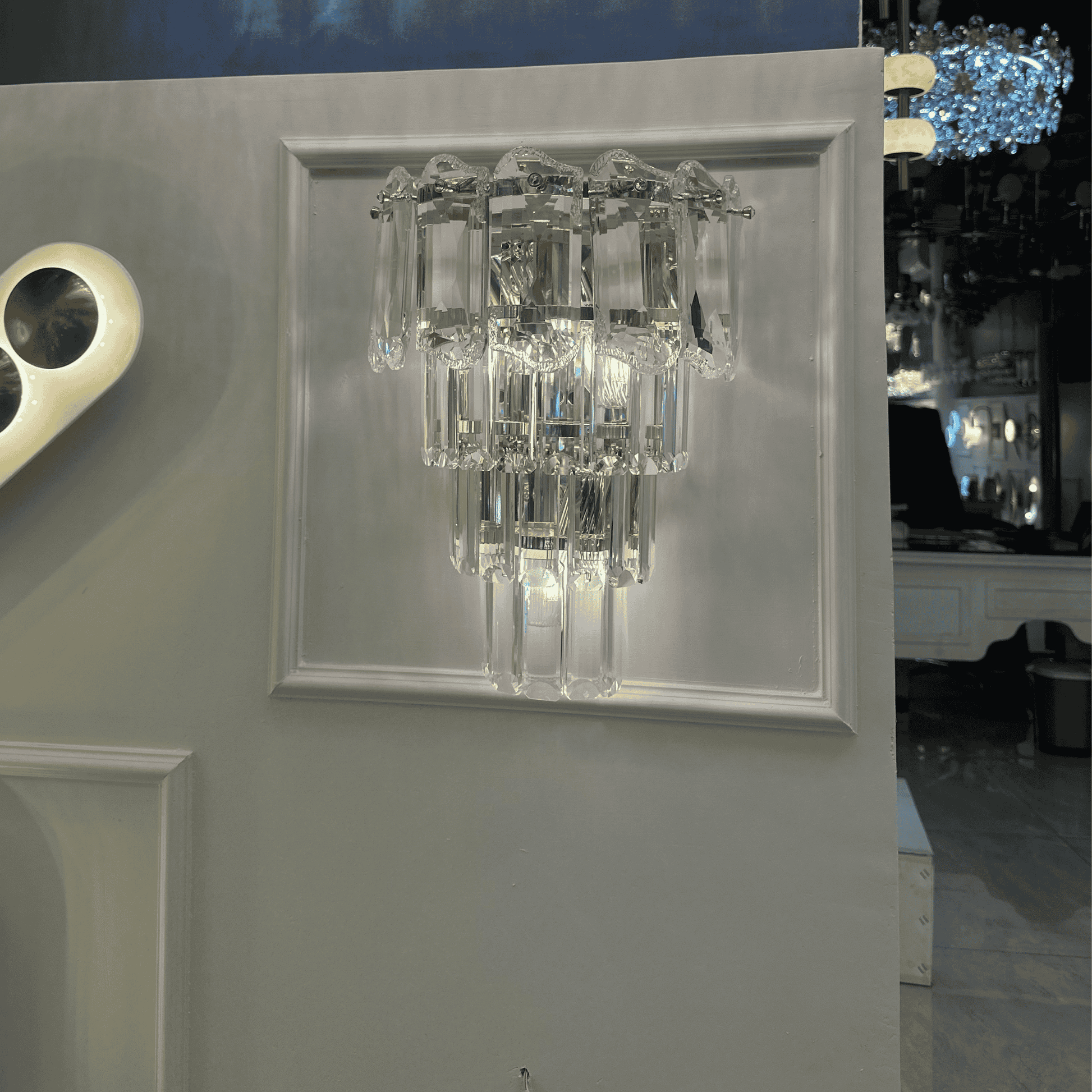 Tiered crystal wall sconce light with elongated clear glass prisms and a chrome frame, mounted on a decorative panel.