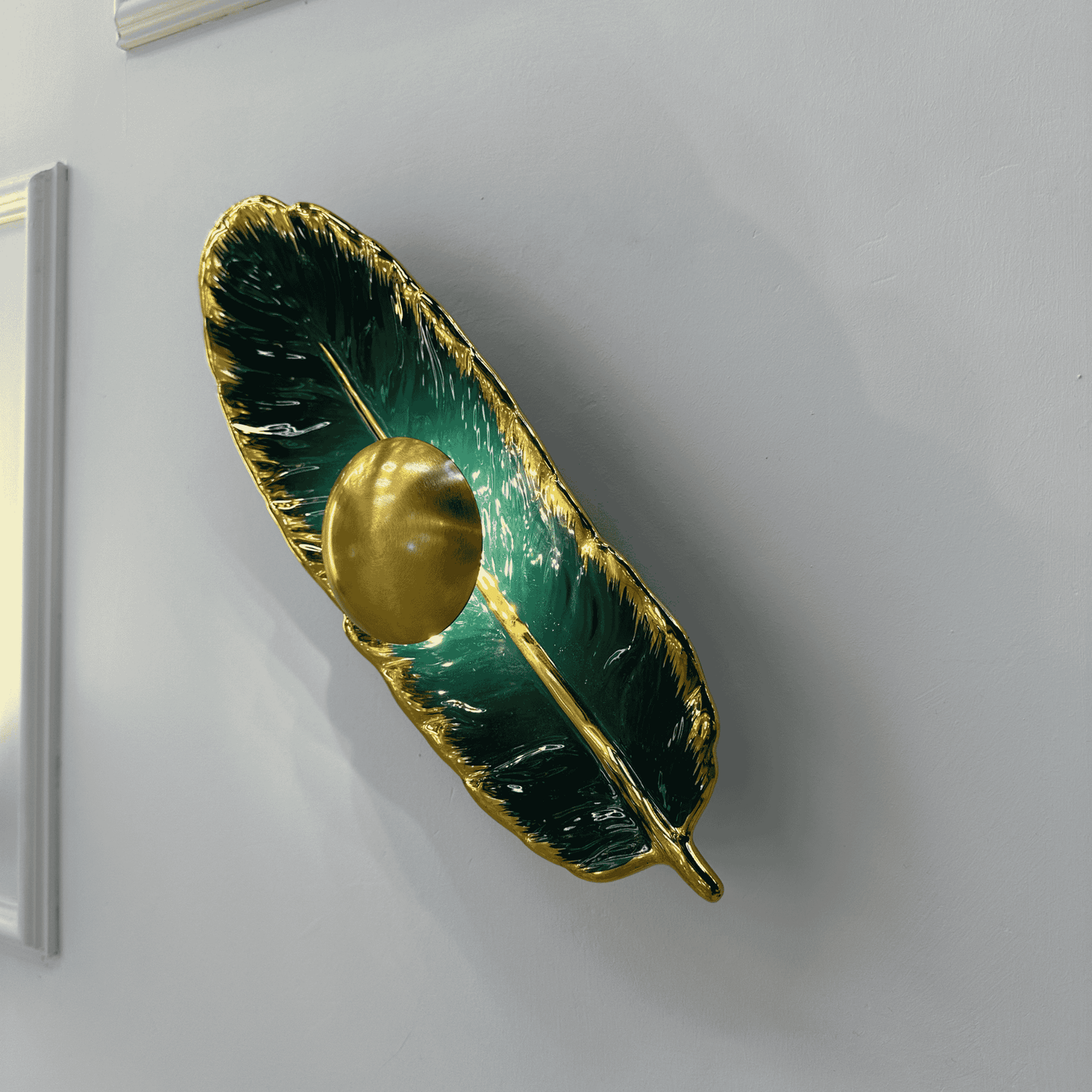 Feather-shaped wall light in green porcelain and brass with LED illumination, providing a decorative modern accent on a plain wall.