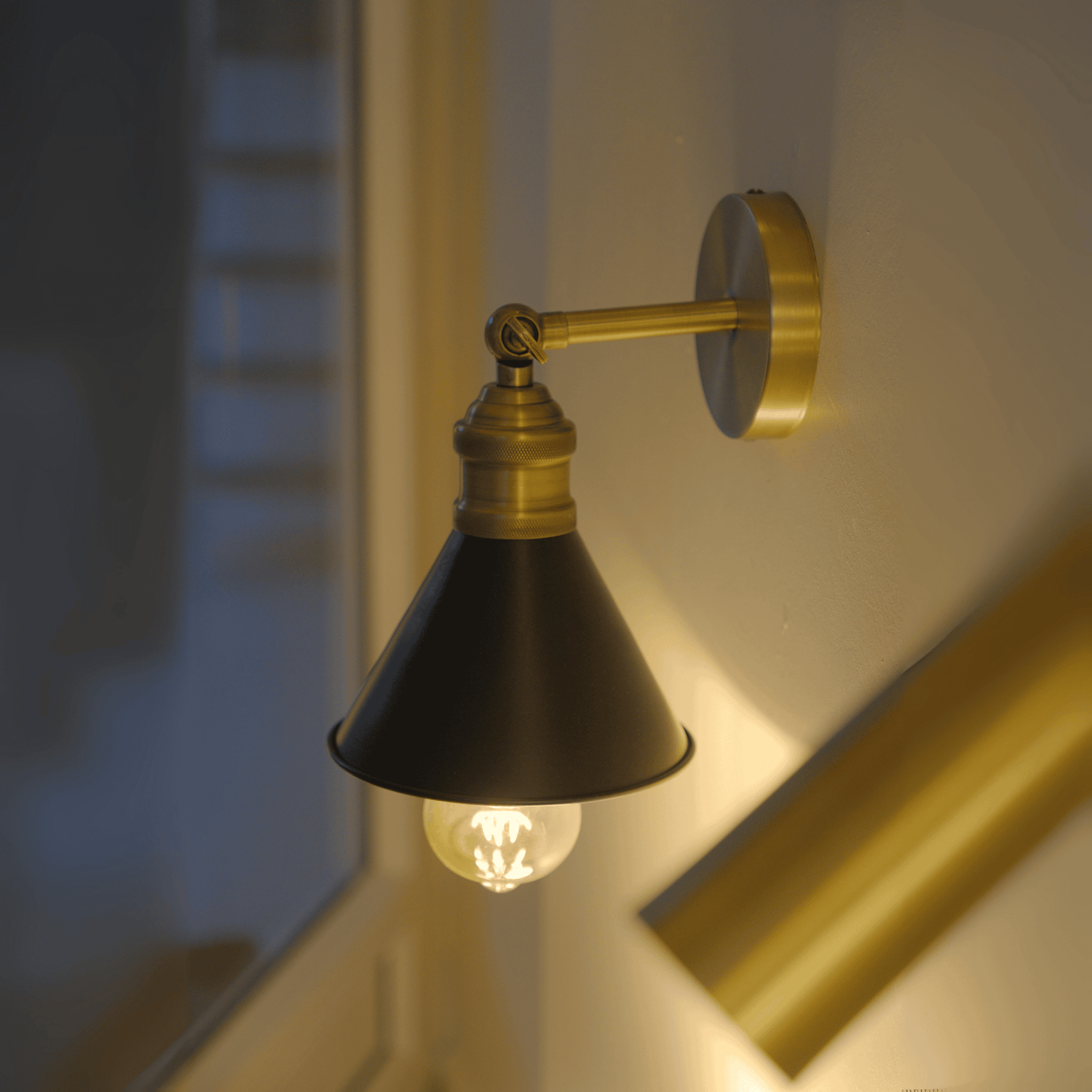 Brass and black wall sconce glowing in a dimly lit interior near a window.