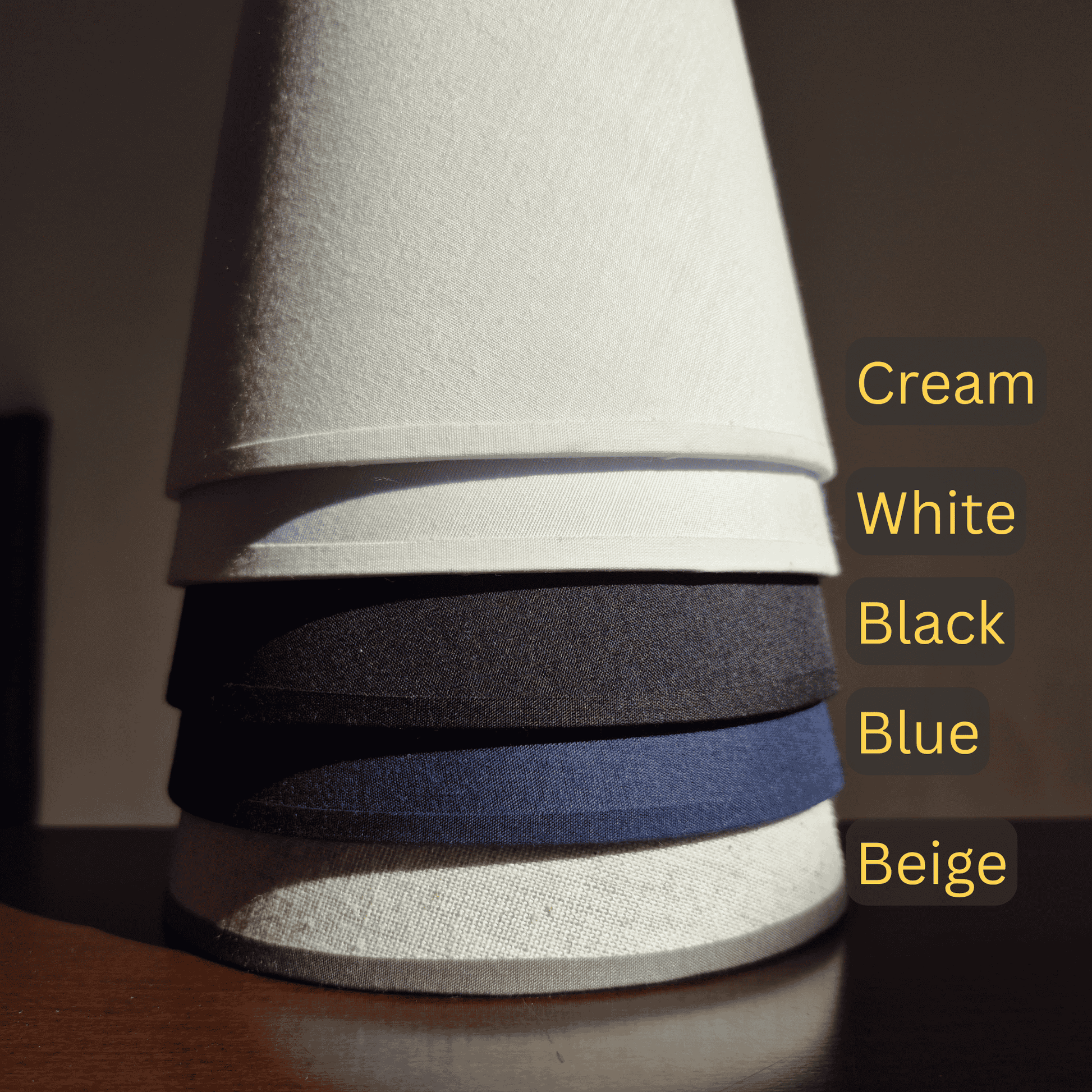 Stack of conical fabric lampshades in five color options: cream, white, black, blue, and beige, labeled clearly
