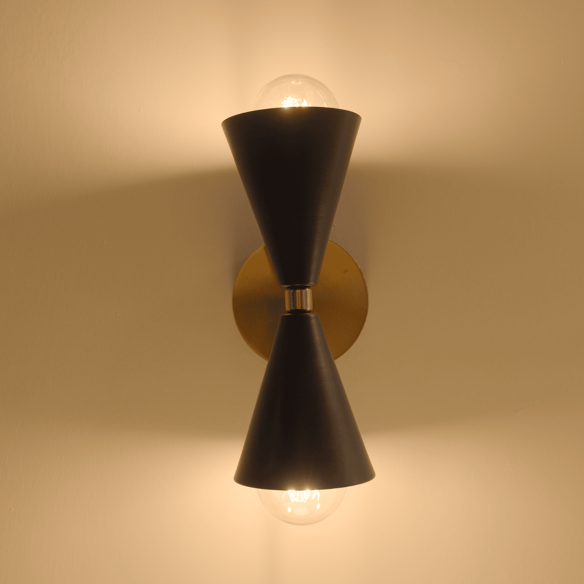 Illuminated black and brass wall sconce with warm lighting casting shadows on a neutral wall.
