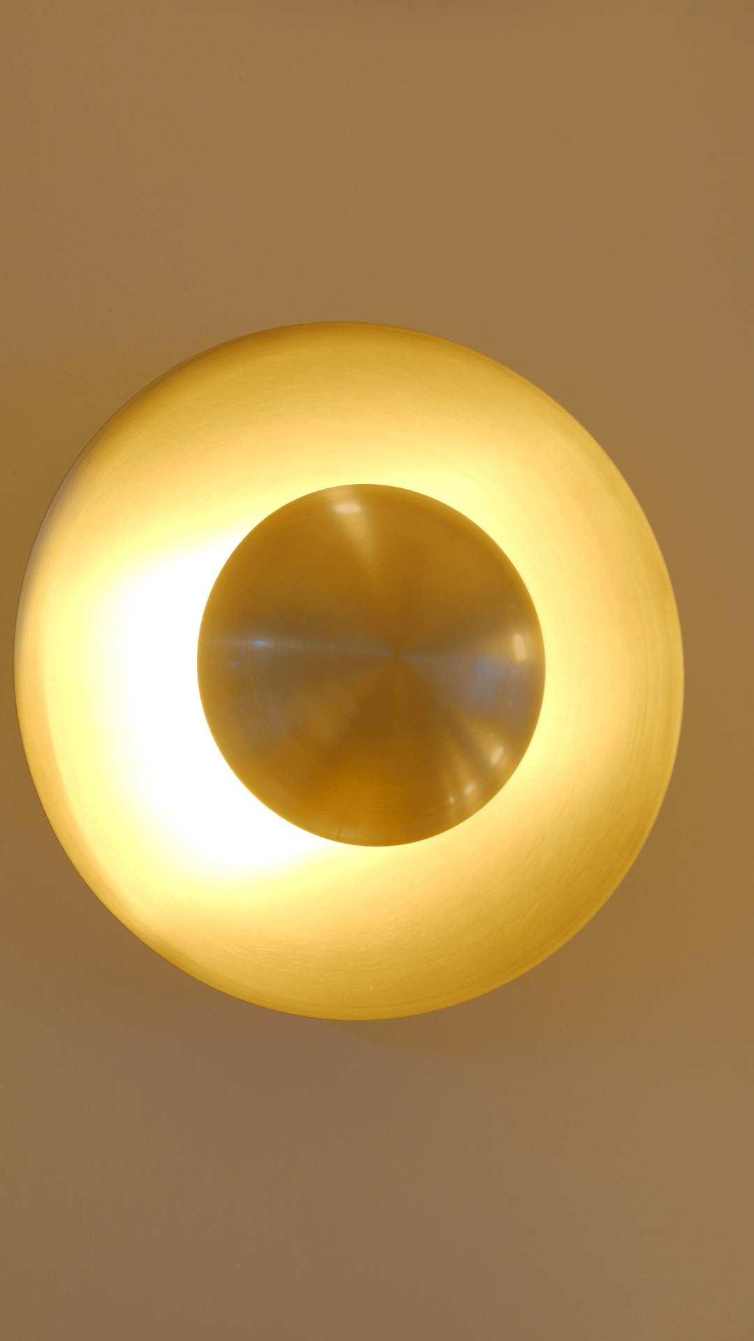 Brass wall sconce with a glowing circular disk emitting warm ambient light on a neutral wall.