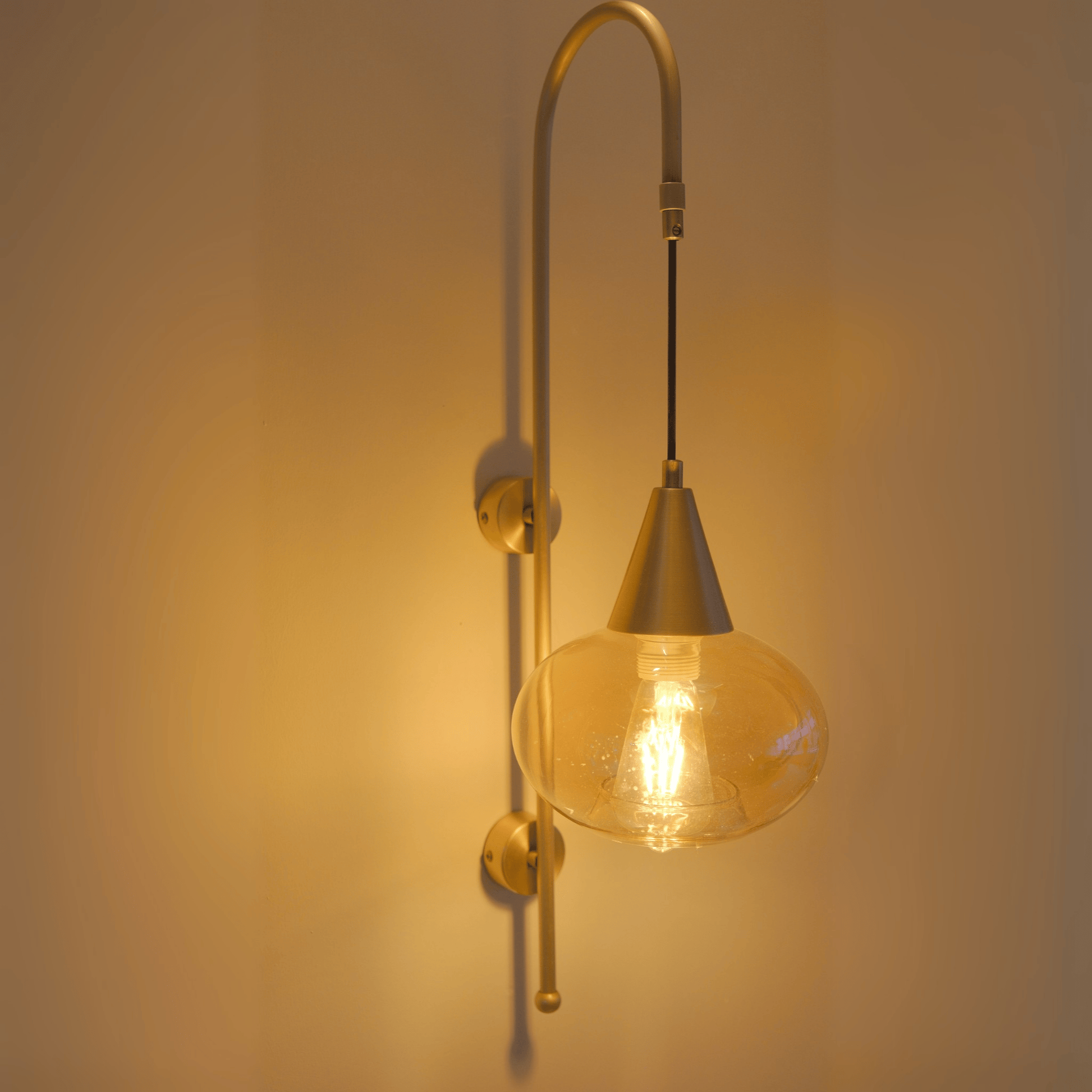 Modern brass wall sconce with a teardrop clear glass shade and exposed filament bulb, emitting soft ambient light.