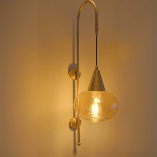 Modern brass wall sconce with a teardrop clear glass shade and exposed filament bulb, emitting soft ambient light.