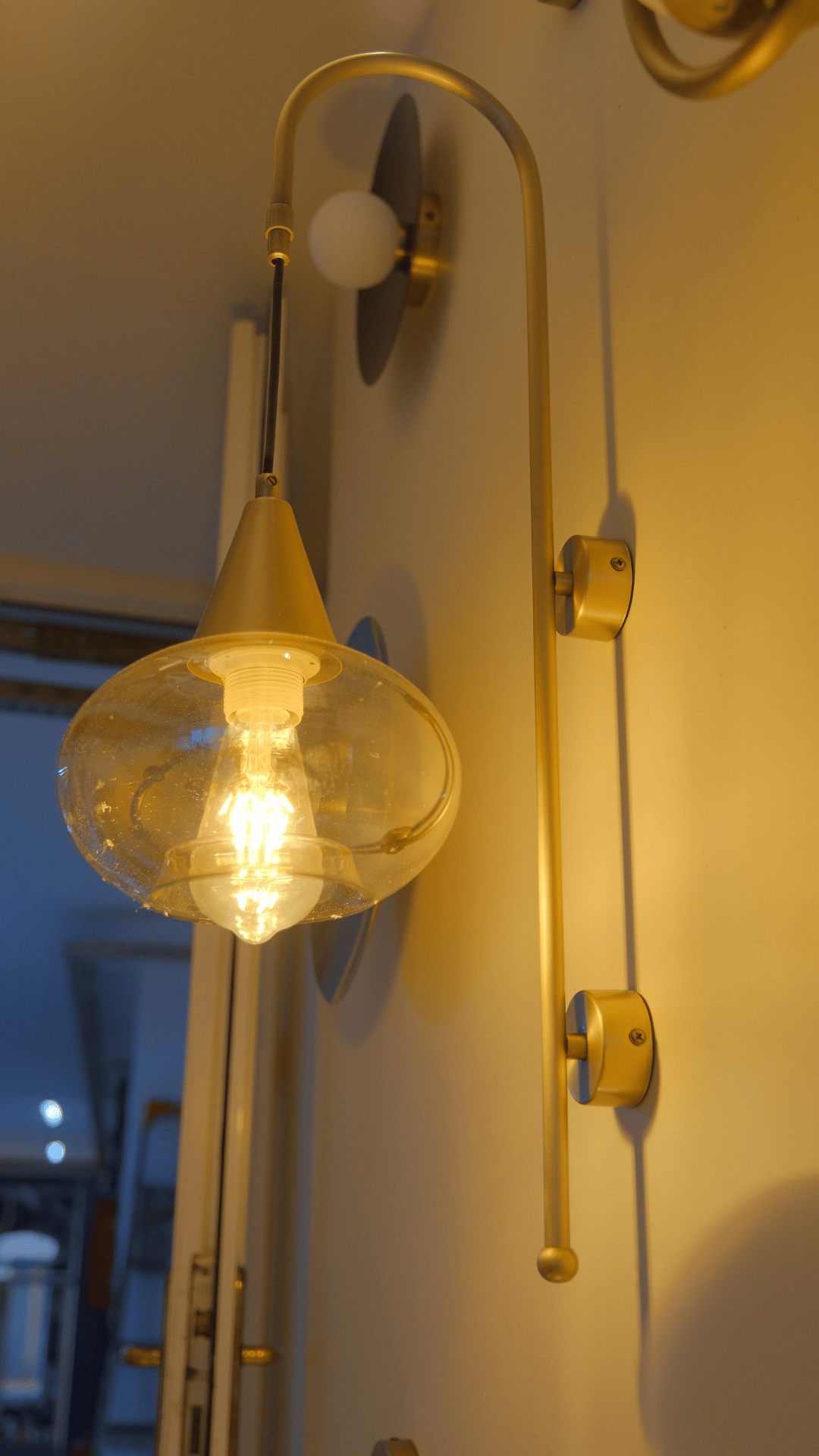 Oversized glass and brass wall sconce with glowing warm LED bulb in hallway.