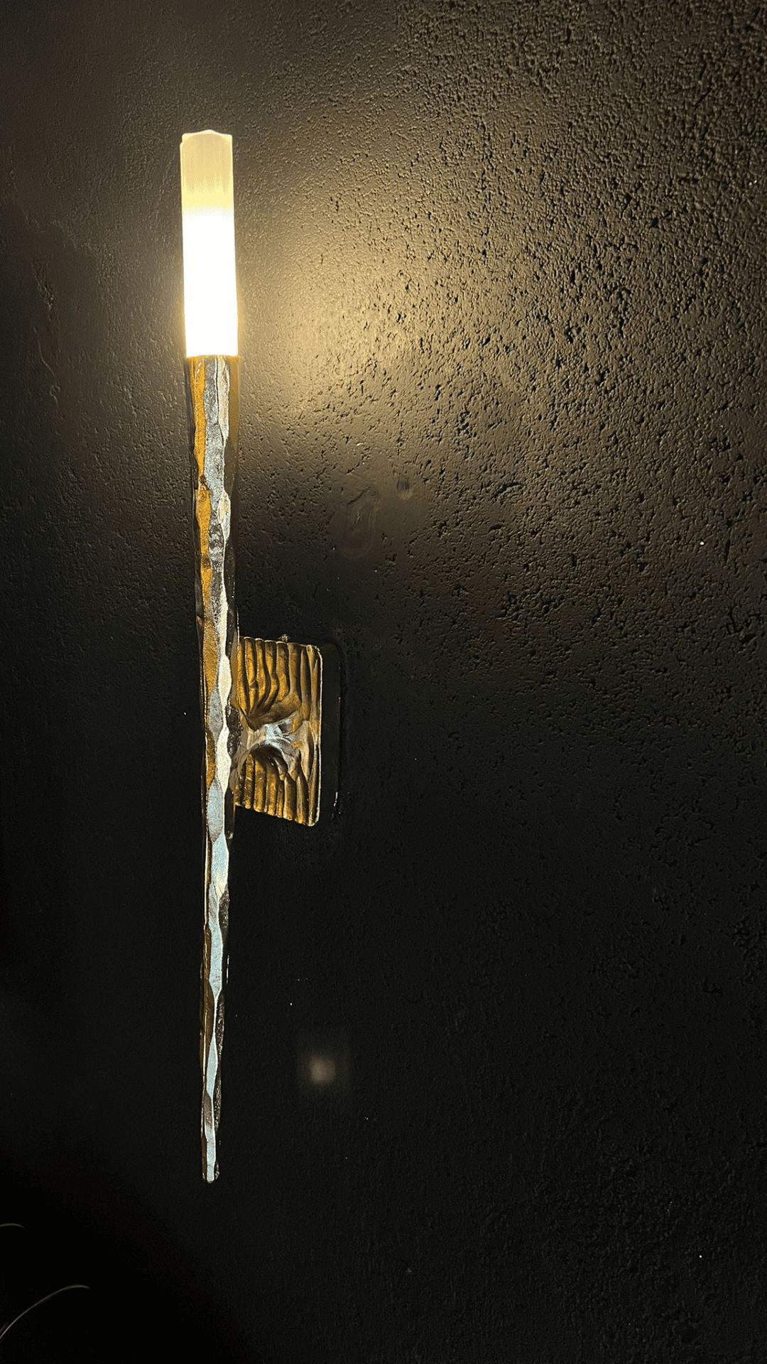 Side view of a hammered brass wall sconce glowing warmly against a dark textured wall.