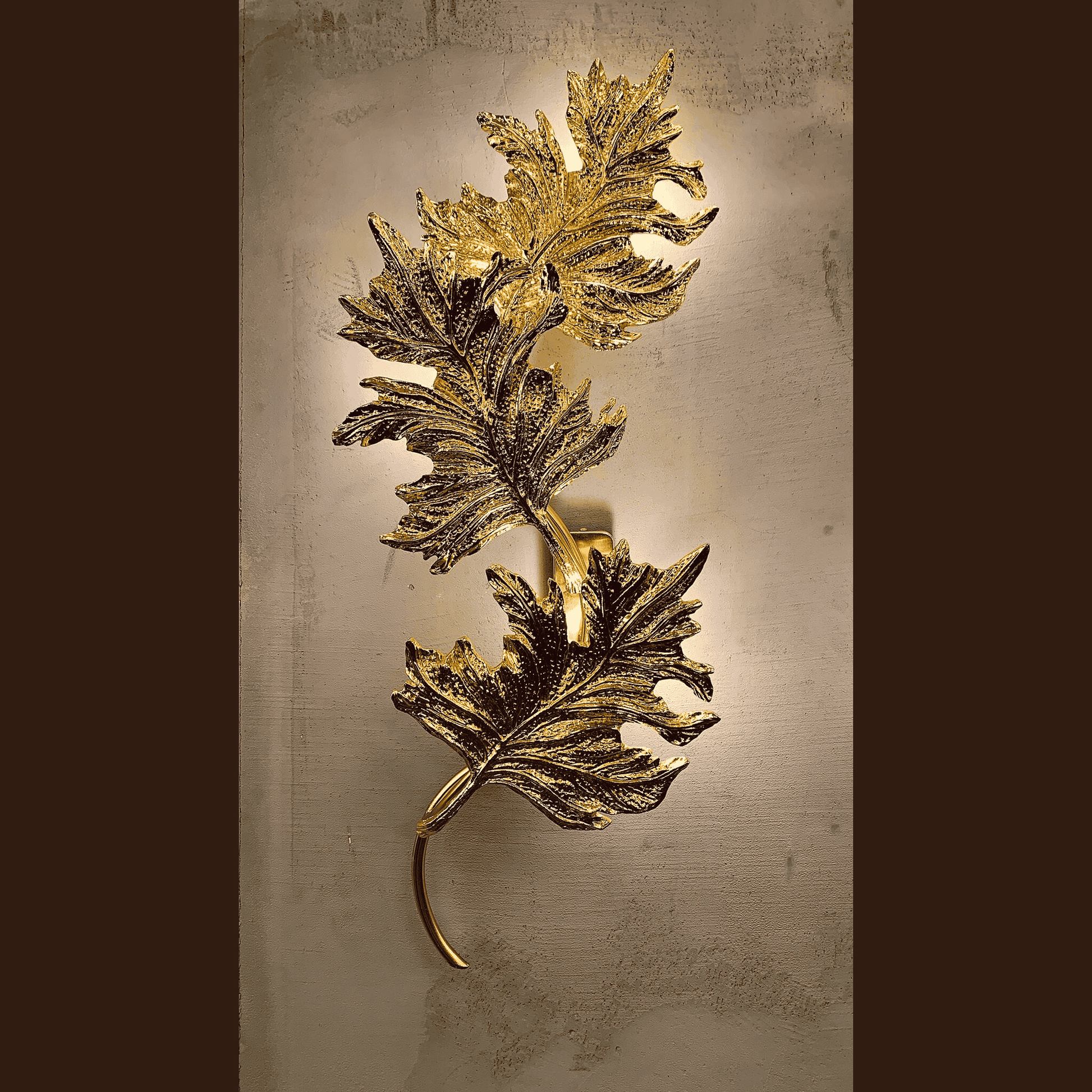 Ornate gold brass wall sconce with sculpted botanical leaf design glowing softly on a textured neutral wall