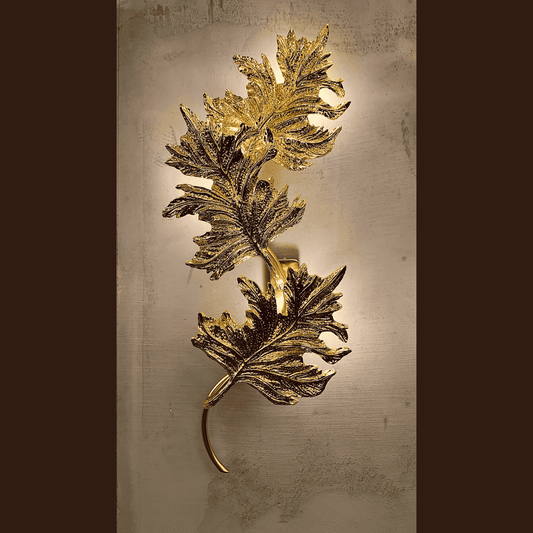Ornate gold brass wall sconce with sculpted botanical leaf design glowing softly on a textured neutral wall
