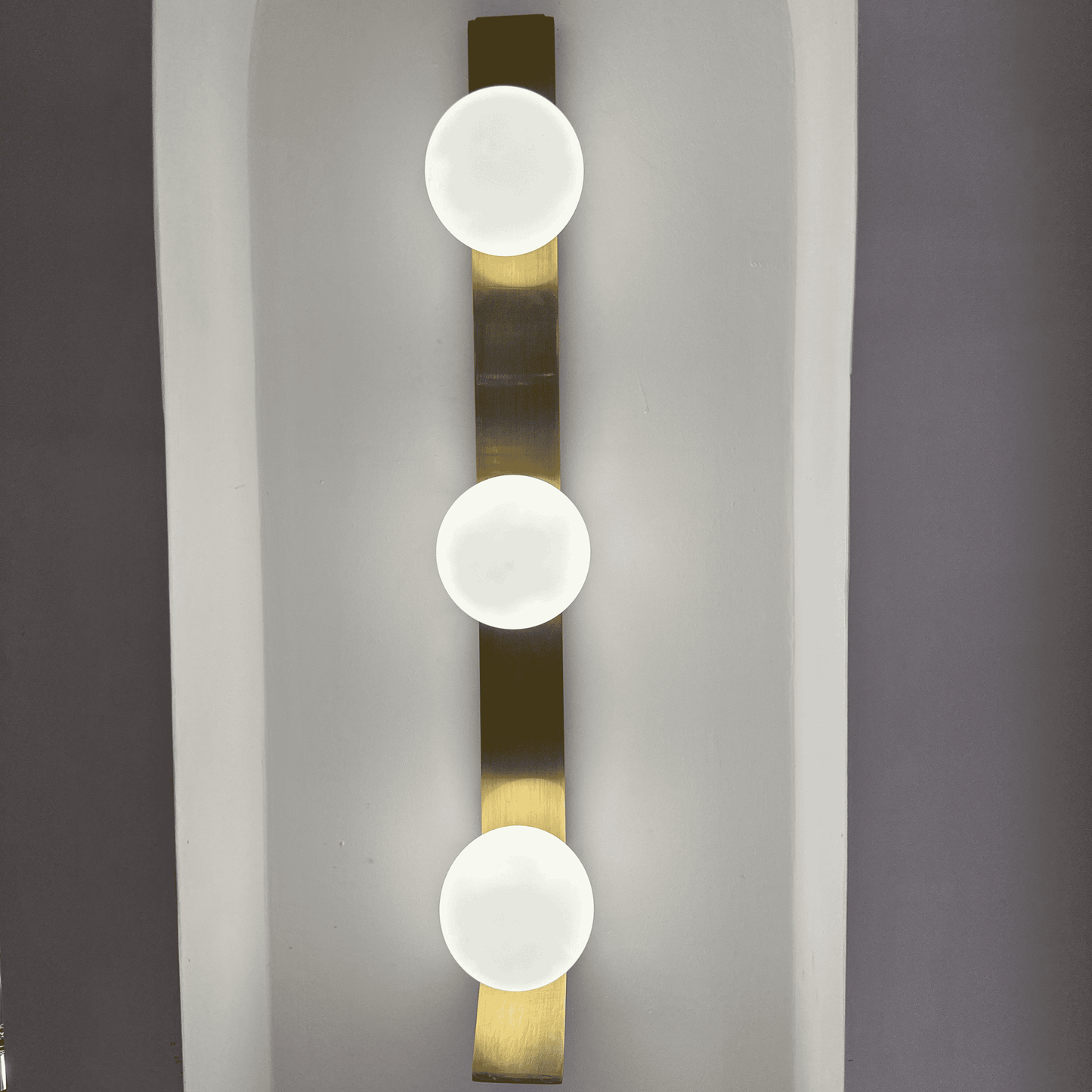 Vertical brass wall sconce with three frosted globe lights mounted on a white wall.