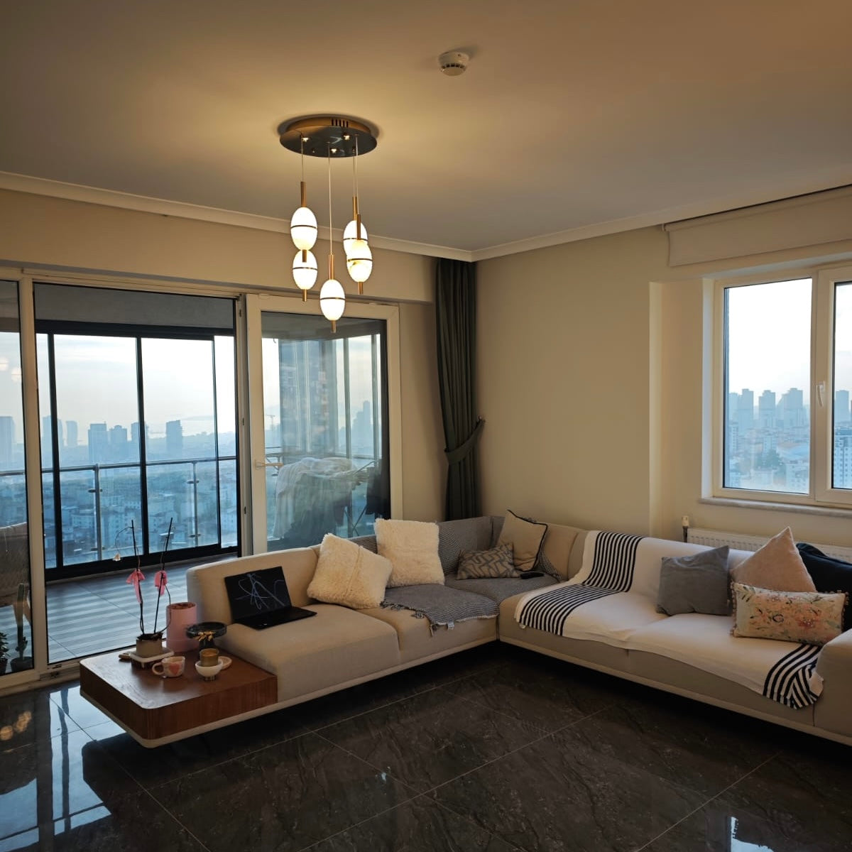 Wide interior view of living and dining area with alabaster pendant lights and city skyline backdrop.