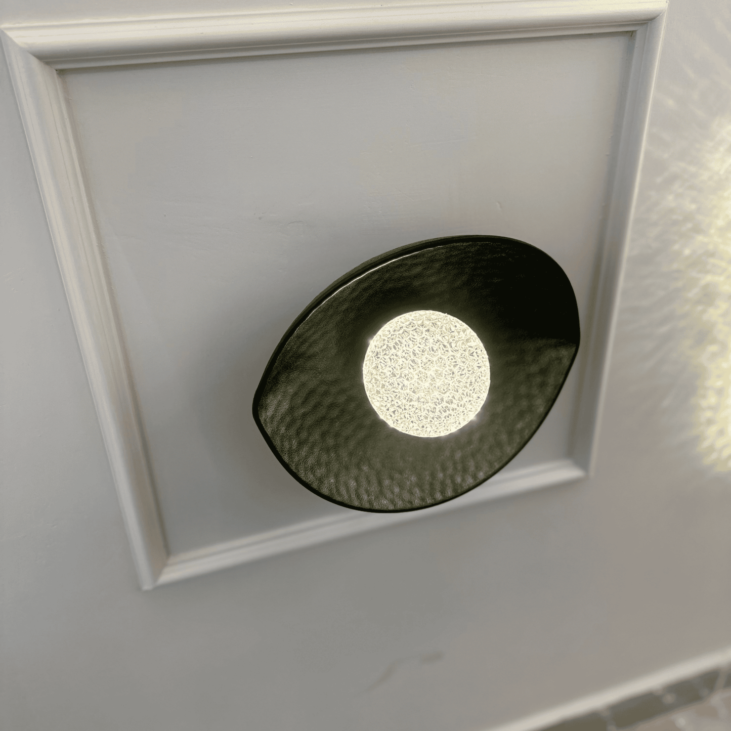 A black plexiglass LED light sconce with a central textured diffuser softly illuminates a neutral-toned wall.
