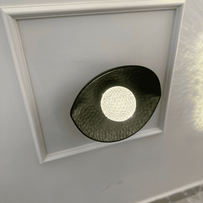 A black plexiglass LED light sconce with a central textured diffuser softly illuminates a neutral-toned wall.