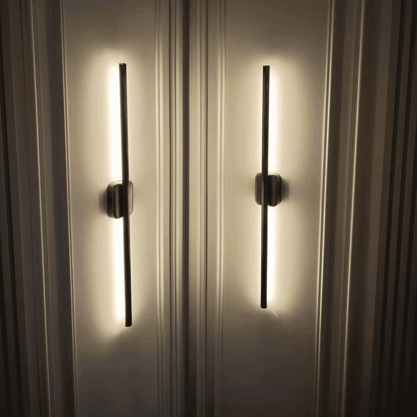 Close-up of two black vertical wall sconces with warm up-and-down light on a decorative paneled wall.