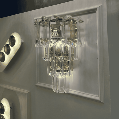 Crystal wall sconce light with layered clear prisms and chrome accents mounted on a wall panel.