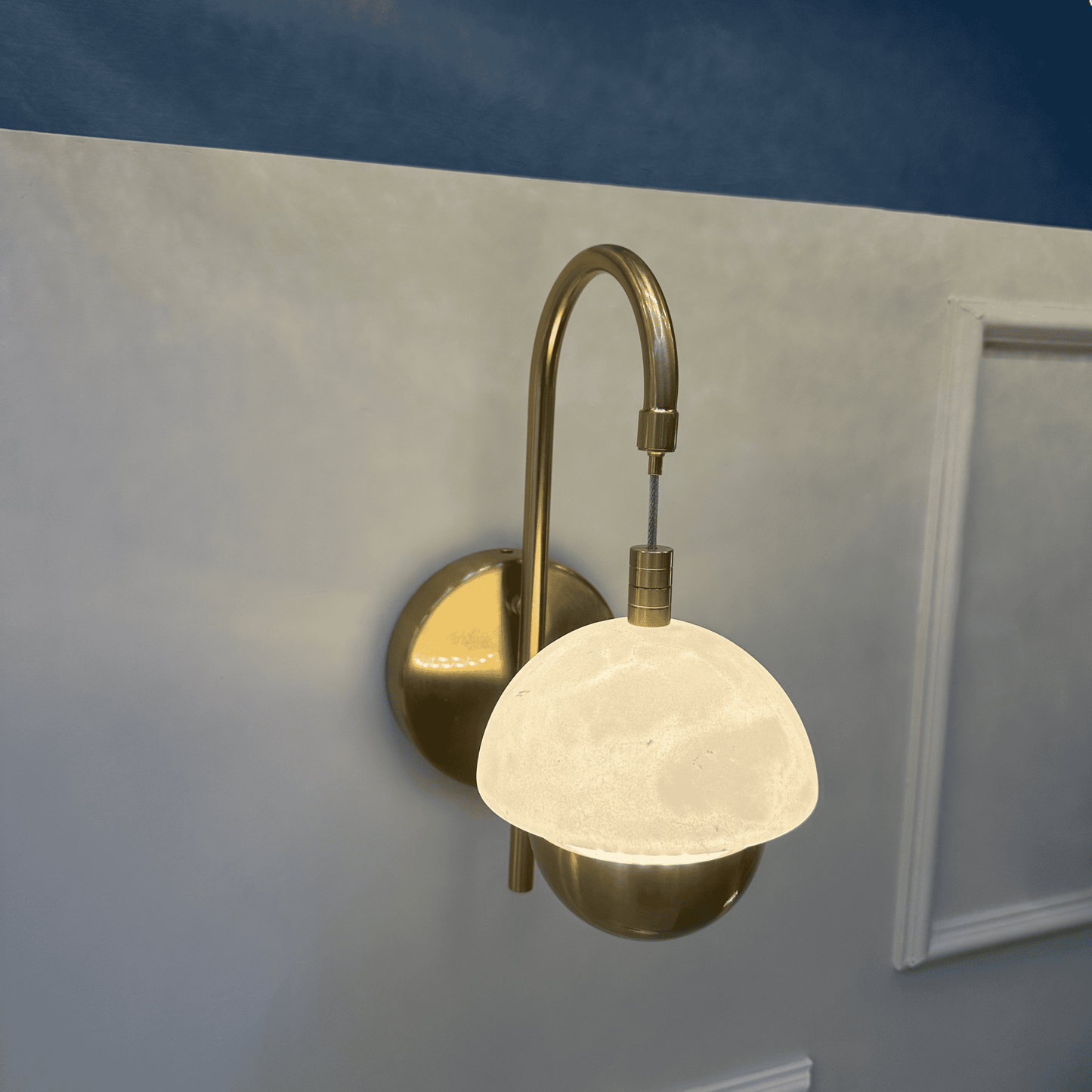 Marble sconce with brass arm glowing warmly on a paneled wall.