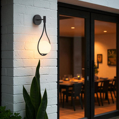 Outdoor modern matte black wall sconce with glowing globe mounted on white brick wall near patio.