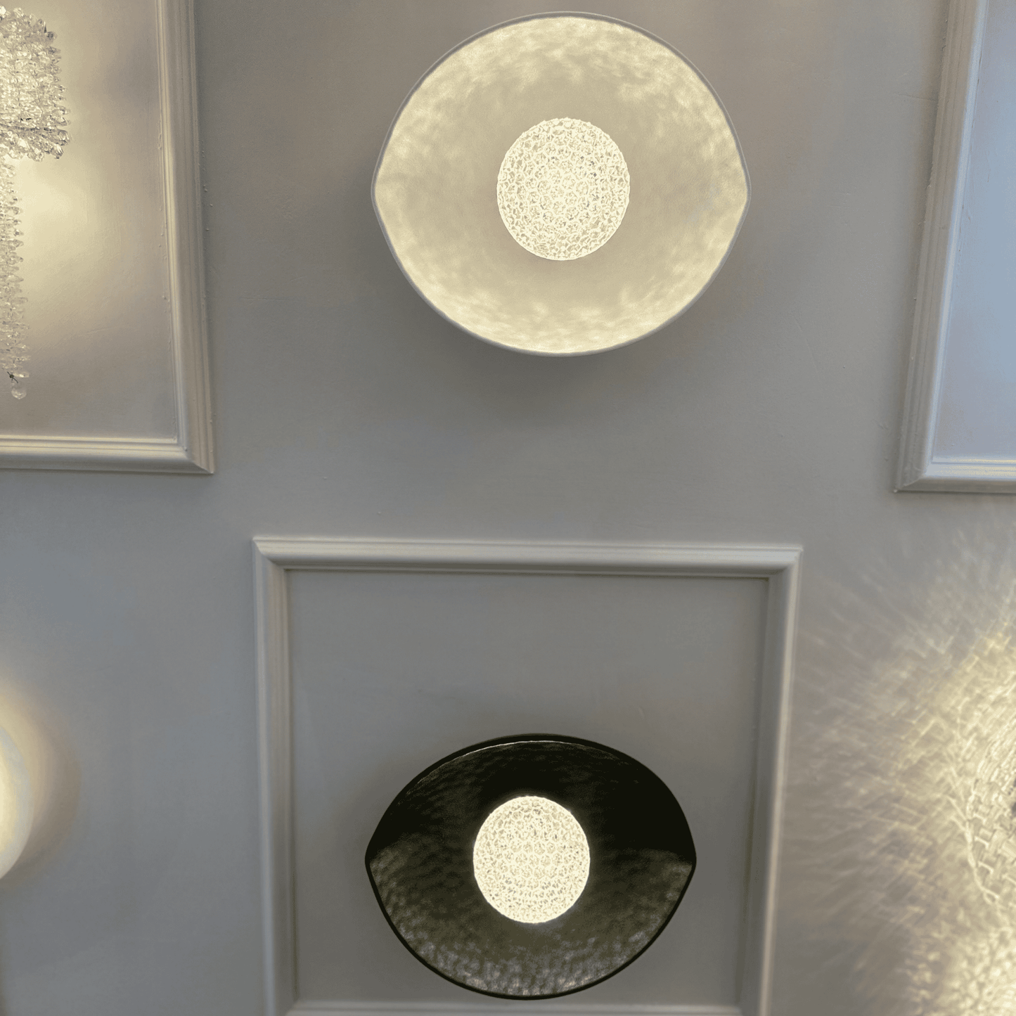 Two plexiglass LED wall sconces in black and white emit soft diffused lighting, mounted on a decorative paneled wall.