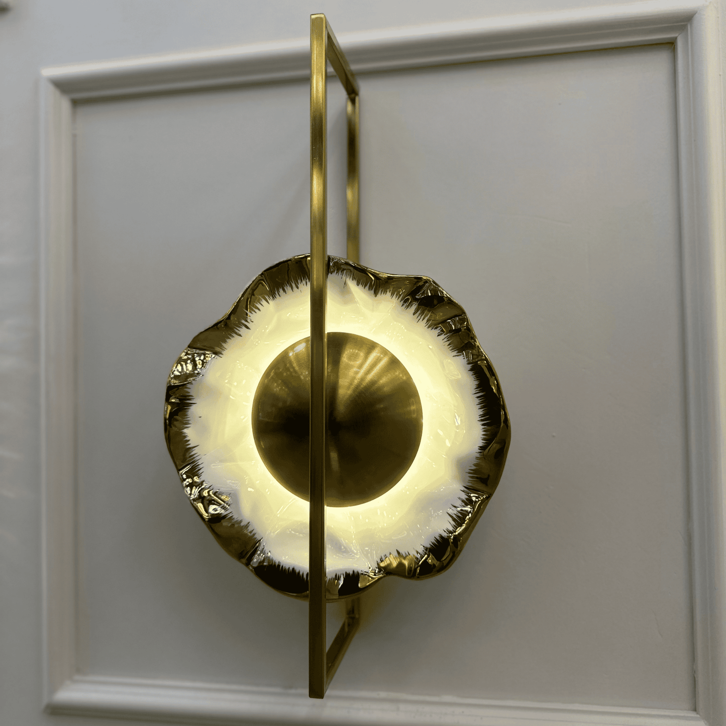 Modern porcelain wall sconce with textured edge and geometric brass frame emitting warm light.