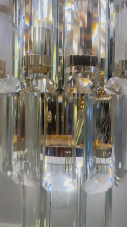 Tiered crystal wall sconce light with elongated clear glass prisms and a chrome frame, mounted on a decorative panel.