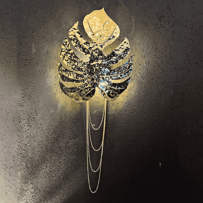 Brass rainforest leaf wall sconce with cascading chain detail mounted on a textured black wall, modern botanical light fixture.