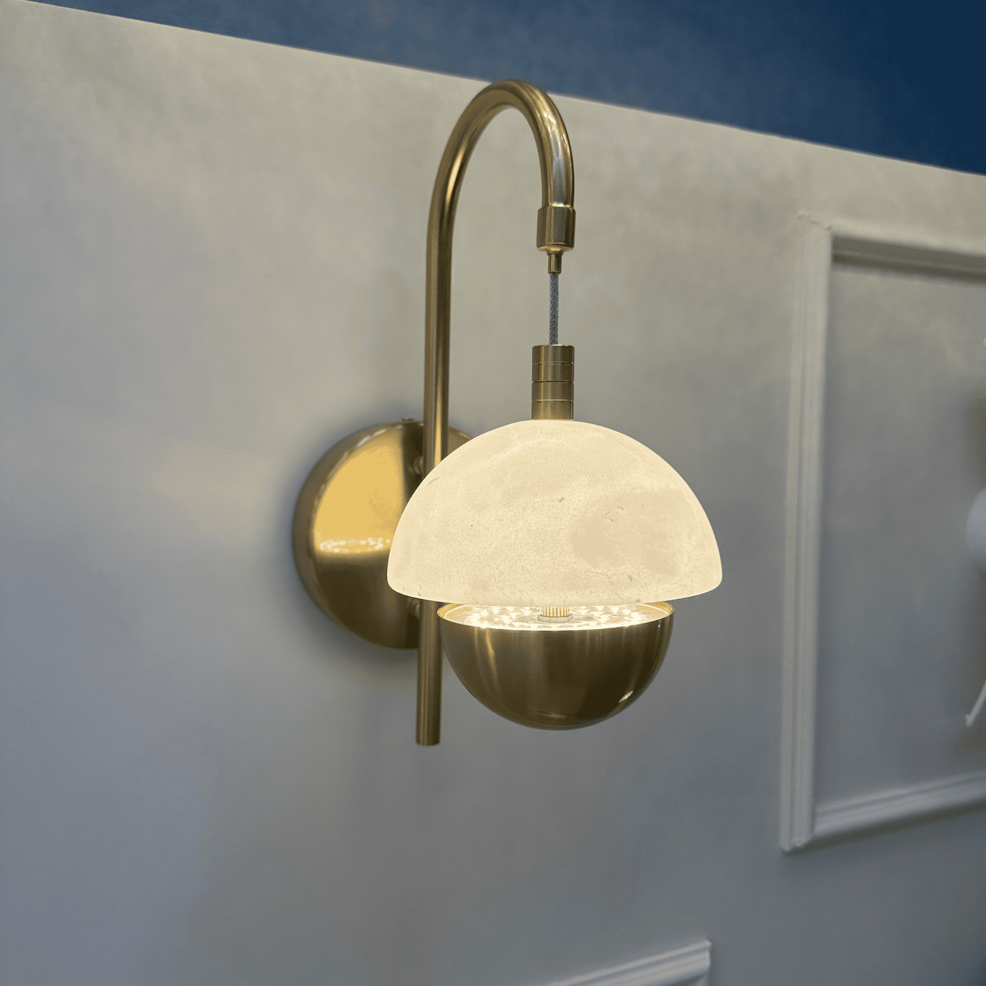 Side view of marble sconce with brass arm and glowing dome diffuser on a light wall.