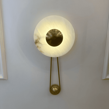 Front view of alabaster wall sconce with central round brass disc on a paneled interior wall.