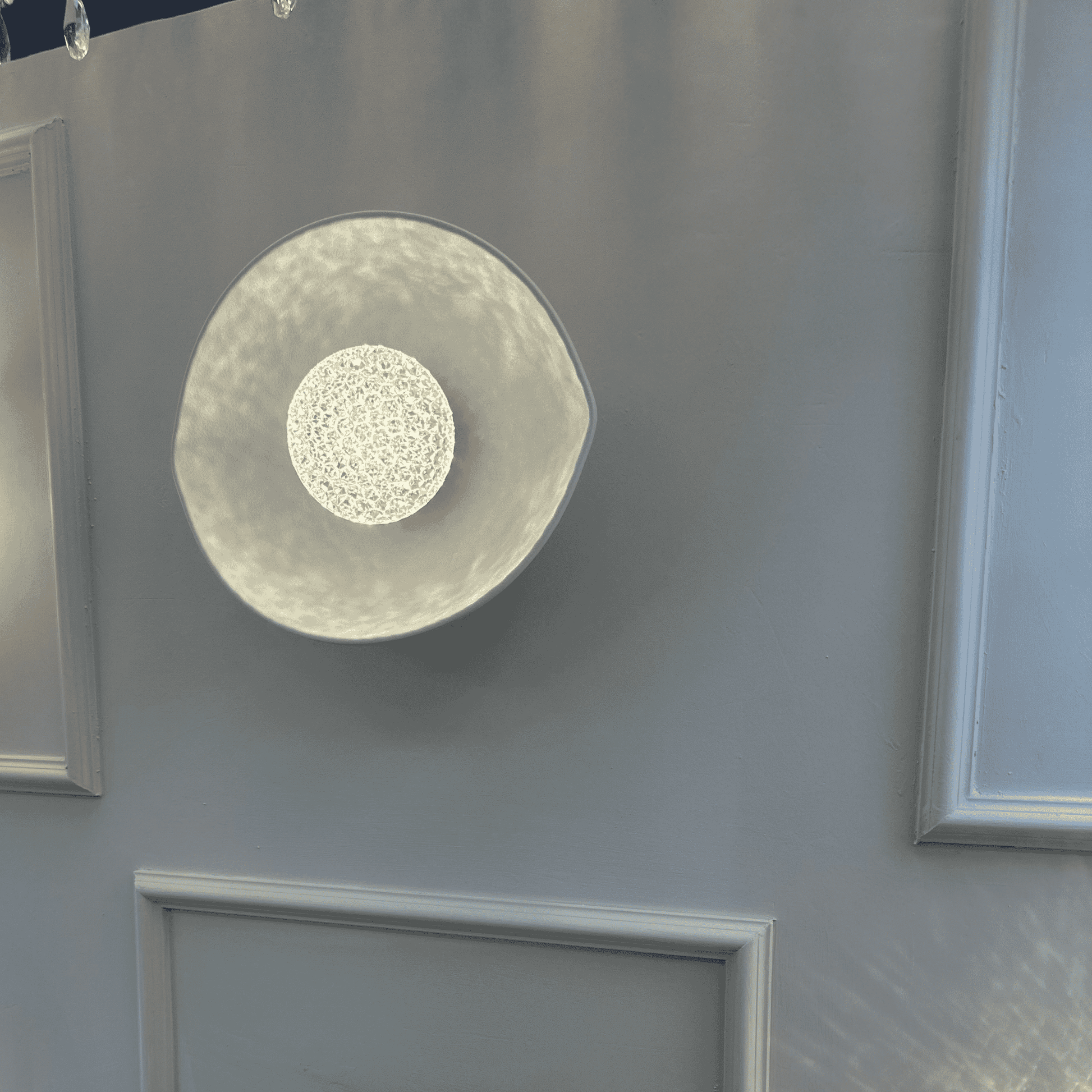 A white plexiglass LED sconce with a textured center emits soft ambient light on a minimalist wall.