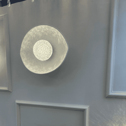 A white plexiglass LED sconce with a textured center emits soft ambient light on a minimalist wall.